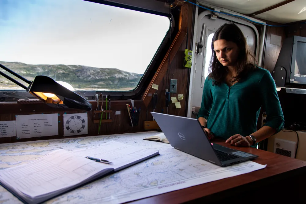 A woman in a ship's cabin working on a laptop at a table.