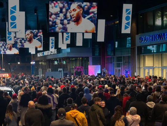 A wide shot of a crowd gathered outside watching large screens that are showing a Toronto Raptors game