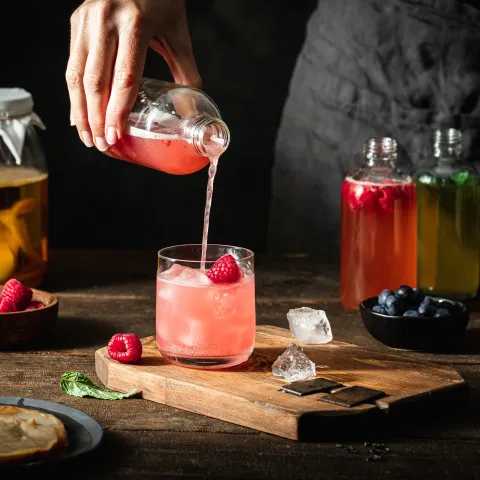 Close-up of woman pouring raspberry kombucha tea in glass from bottle. Female serving homemade flavoured fermented tea.