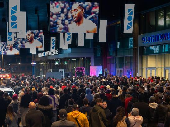 A wide shot of a crowd gathered outside watching large screens that are showing a Toronto Raptors game