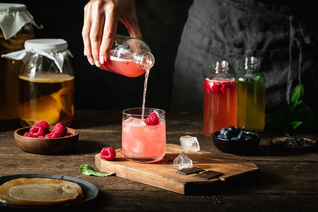 Close-up of woman pouring raspberry kombucha tea in glass from bottle. Female serving homemade flavoured fermented tea.